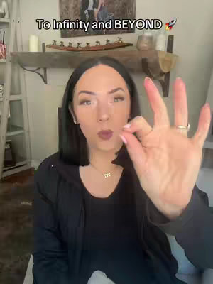 A woman with dark hair is smiling and giving a thumbs-up. The text 'To Infinity and BEYOND' is at the top of the frame, along with a rocket ship emoji. She is wearing dark clothes and a necklace. The background is a room with a fireplace and decorative items.