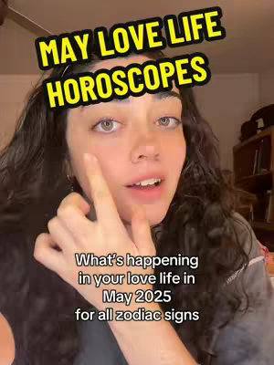 A woman with long dark hair and fair skin is centered in the frame with a soft, indoor background. She is looking towards the right while pointing towards her cheek with her right index finger. Above her, a rounded rectangle, yellow, overlay says "MAY LOVE LIFE HOROSCOPES" with a black stroke. Below her, there is a white text that says "What's happening in your love life in May 2025 for all zodiac signs".