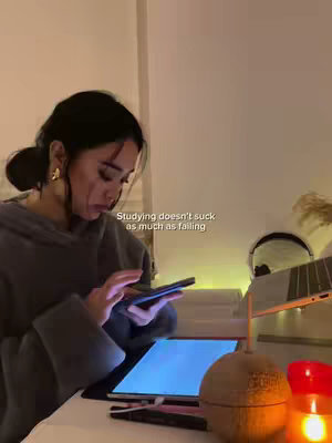 A woman is seated at a desk, illuminated by warm lighting. She is interacting with a phone, while a tablet is on the desk, which features a bright, blue screen. Additional elements include a candle, a desk lamp, and objects that create a cozy atmosphere.
