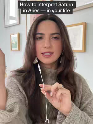 A woman with brown hair and a beige sweater speaks into the camera, holding a white earphone cord. The background shows a bright room with framed pictures and a wall painted light green. The text overlay reads 'How to interpret Saturn in Aries — in your life'.