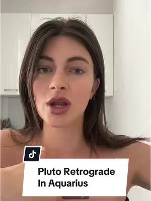 A woman is centered in the frame, looking directly at the viewer with a serious expression. Behind her are white cabinets. A white rectangle at the bottom with the title "Pluto Retrograde In Aquarius".