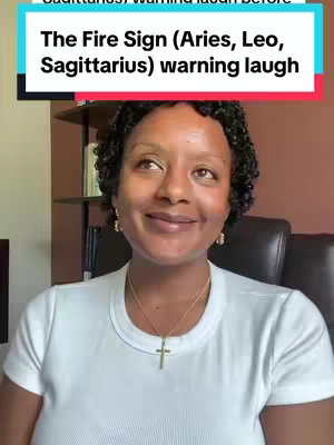 A woman with dark curly hair, wearing a white t-shirt and a gold cross pendant, looks up and to the left with a slight smile. Above her is a text overlay in a teal box that reads 'The Fire Sign (Aries, Leo, Sagittarius) warning laugh'. The background is a room with a bookshelf and dark chair.