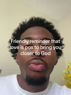 A man of color with short curly hair is featured in a selfie-style shot. The background is a plain, light-yellow wall, and there are hints of a yellow flower in a vase to the bottom right. The man is wearing a plain white shirt and has text overlays. His mouth is open and his tongue is protruding.