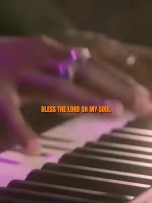 The image shows a close-up of a person's hands playing a piano. The keys are black and white, with the hands positioned to play a chord. Above the keys, the text "BLESS THE LORD OH MY SOUL" is written in an orange font, with blurred background that suggests a performance setting with dim lighting and colorful light effects.