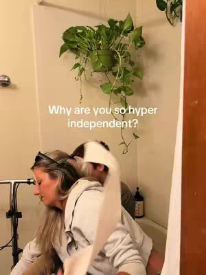 A woman is shown from the side, sitting in front of a bathtub. A roll of toilet paper is being held against her back as she turns her head toward the side. The background includes a Pothos plant hanging on the wall.