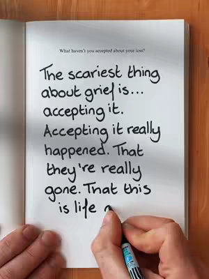 The image shows an open book on a wooden surface, with handwriting on a white page. The text, in black ink, begins with the phrase, "The scariest thing about grief is..." A hand, holding a black pen, is visible at the bottom, seemingly in the process of writing or underlining the words.