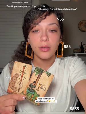 A woman with curly hair and large gold hoop earrings holds two tarot cards in front of her. Text overlays on the image include numbers and phrases like 'New Moon in Cancer'and  'Booking an unexpected trip'.  The woman is centered, looking towards the viewer with a neutral expression.