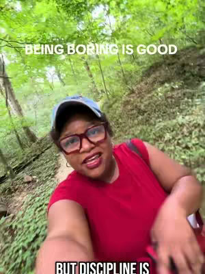 A selfie-style shot taken from a slightly low angle, with an individual in the foreground. The person is wearing glasses, a baseball cap, and a red shirt. The background consists of dense green foliage, suggesting a forest or park setting, with text overlays.