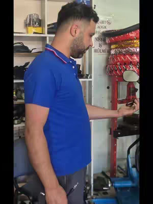 A man with a beard stands in a garage or workshop, holding a tire tool while facing a red and blue tire-changing machine. Behind him, there are shelves with tools and tires.