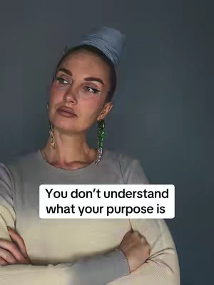 A woman with a serious expression is the focal point. She has her arms crossed. Above the woman is a background of a solid grey color. The woman is wearing a light grey shirt. She has a head wrap and unique earrings. A text overlay in a white box states, "You don't understand what your purpose is".
