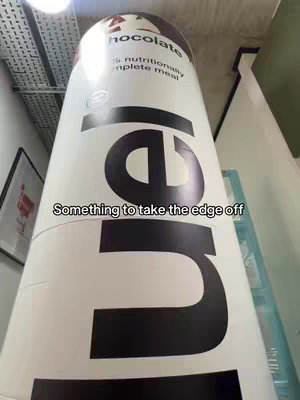 The image shows a large, cylindrical object that appears to be a product display, likely a complete meal replacement by Huel. The structure is predominantly white and features large black text and the Huel logo. The word 'Chocolate' is visible at the top, indicating the flavor. Text overlayed at the bottom states: 'Something to take the edge off.' The background includes parts of a ceiling and other architectural elements, suggesting an interior setting. The lighting appears to be from above, creating highlights on the cylindrical surface.