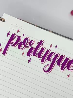 The image shows a close-up of a notebook page with the word "português" hand-lettered in large, cursive purple letters. The letters are outlined in a darker shade of purple, and there are small diamond-shaped decorations around the word. The background is partially visible, showing a spiral binding and patterned paper.