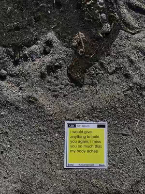 The image shows a close-up view of dark, granular sand. A small, yellow sticky note is resting on the sand, its surface covered with dark text. The background consists of the sand, which takes up most of the frame, textured with tiny, dark grains. The note has the remnants of screen elements found on mobile phones or tablets.