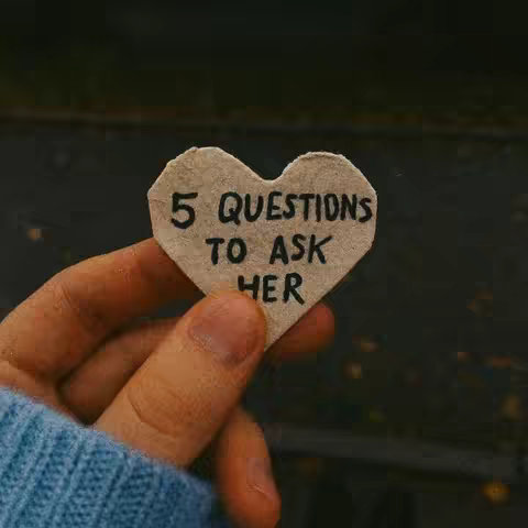 A hand holds a heart-shaped piece of paper with the text overlay '5 QUESTIONS TO ASK HER'. The background is out of focus, suggesting an outdoor setting. The heart-shaped paper is light brown. The hand is holding the paper, and part of a blue sleeve is visible.