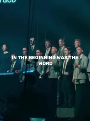 A group of men in suits are standing on a stage holding microphones. Text overlayed on the image reads, "IN THE BEGINNING WAS THE WORD". The background is dark and out of focus with some white text at the upper area of the image.