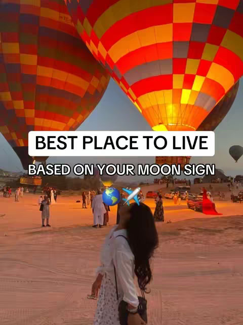 A person stands in front of two hot air balloons, one red and yellow and the other orange and red with yellow and white. The person is wearing a white top and patterned skirt. The sky is clear, and there are many people and some cars in the background. On the image are text overlays in a white rectangle.