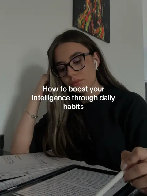 A young woman with long brown hair and glasses is seated at a desk with books and a tablet. She is wearing earbuds and a black shirt, and is appearing to be studying. The image has text overlayed in the center that states, "How to boost your intelligence through daily habits."