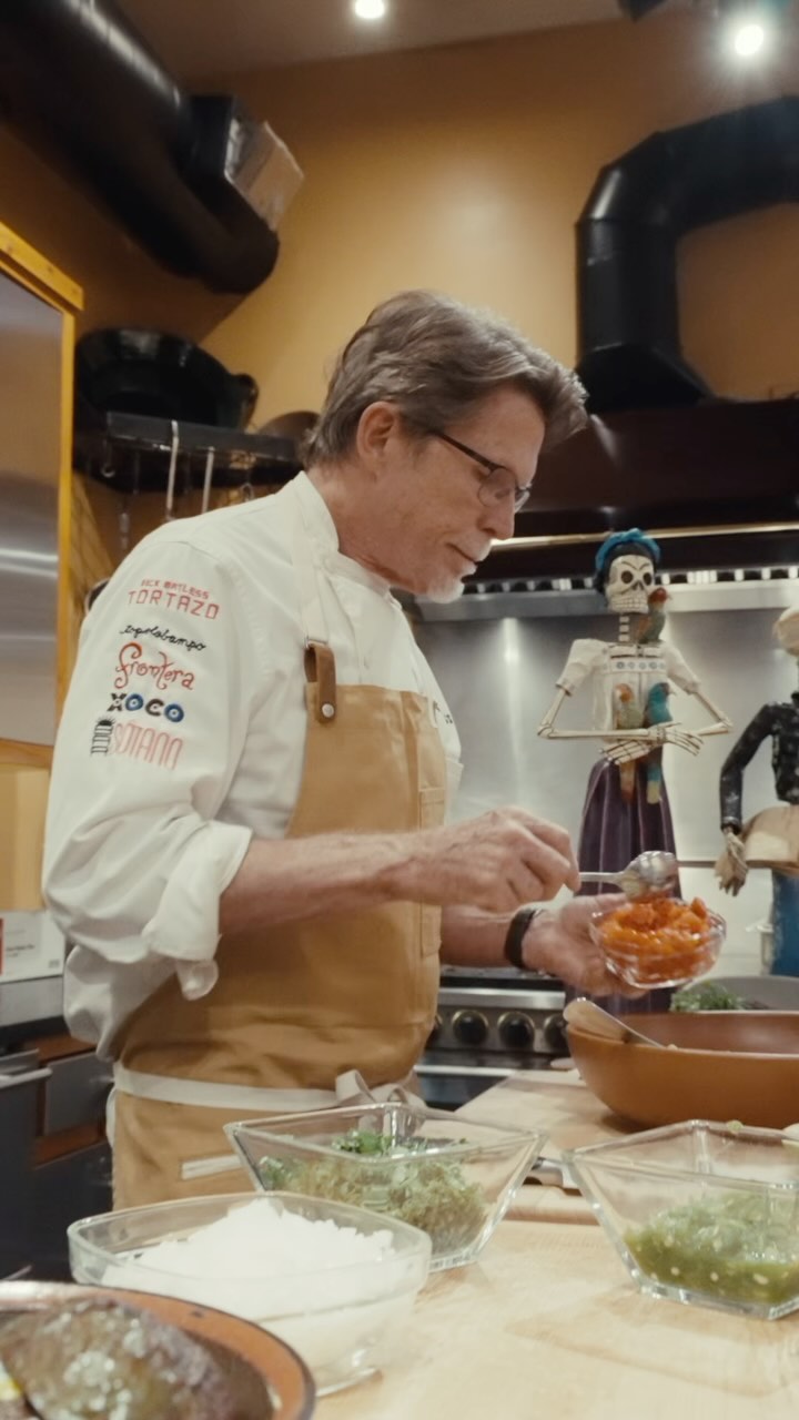 A man in a chef's coat with a beige apron is preparing food, holding a small bowl of chopped red tomatoes in his right hand and a spoon in his left, with various ingredients in glass bowls in front of him. The background includes a kitchen setting with decorations.