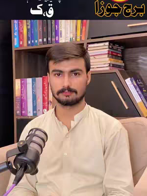 A man with a mustache is centered in the frame. Behind him is a bookshelf and a desk with a microphone in front of him. He is wearing a light-colored shirt and sits in front of a tan couch.