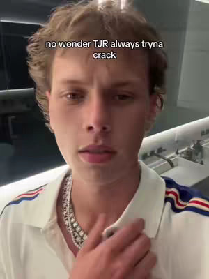 A young man with wavy light brown hair is centered in the frame, gazing directly at the viewer with a slightly concerned expression. He's wearing a white polo shirt with blue, red, and white stripes on the sleeves. A silver chain necklace sits around his neck. The background seems to be a blurred interior with indistinct elements.