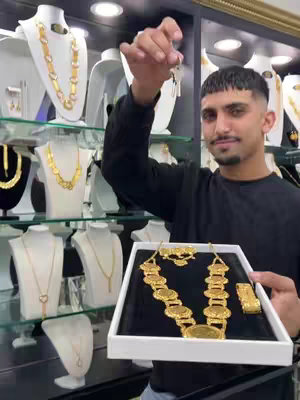A man stands in a jewelry store, holding a tray of gold jewelry. Behind him, various necklaces are displayed on mannequins, and the store is lit by overhead lights. The man holds keys in his other hand, and has a slight smile on his face.
