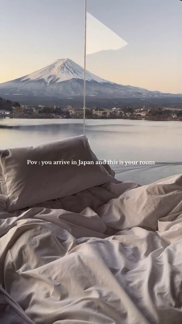 A view from a bed inside a hotel room in Japan. The scene is looking out of a floor to ceiling window at snow-capped Mount Fuji in the distance. The foreground features rumpled gray sheets and a pillow from the bed. The light is soft, creating a calm and serene atmosphere.