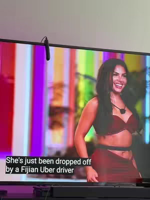 The image captures a scene from a television screen. The screen shows a woman in a red outfit with a necklace smiling. Overlaid on the screen is a text box with the words 'She's just been dropped off by a Fijian Uber driver'.