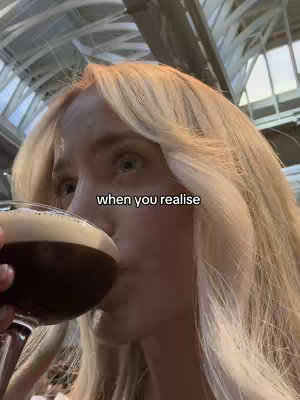A selfie-style image featuring a woman taking a drink from a cocktail glass. The image has a blurred background with overhead lighting.