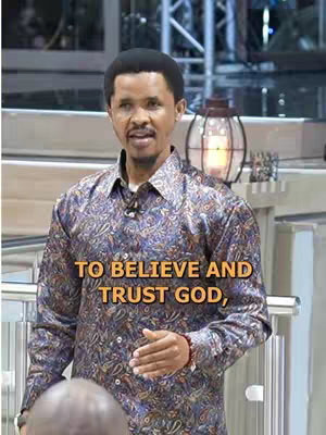 The image captures a medium shot of a man in a patterned blue and brown button-up shirt addressing a live audience. The background is a softly lit interior space with visible metal railings, a decorative candle lantern, and lights. The man has dark skin and short, well-groomed hair. Overlaid text reads, "TO BELIEVE AND TRUST GOD". 