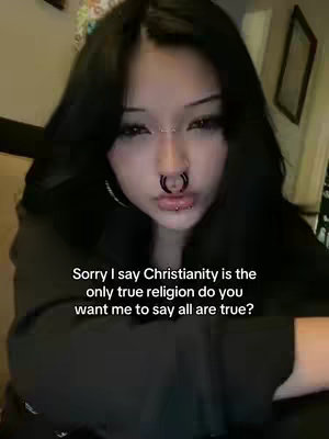 A selfie-style shot featuring a person with long dark hair in front of what appears to be an indoor setting. The person has multiple facial piercings, including a nose ring. Text overlayed on the image contains text about a religious claim.