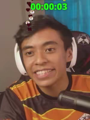The cover image shows a young man in a black and orange sports jersey, smiling directly at the camera. He's positioned centrally, with parts of a microphone visible in the bottom right, and a headset around his neck. Above his head and slightly left, floating elements of hearts and profile icons are displayed. Behind him is what appears to be a gaming chair, with a blurry background hinting at a room. The overall impression is of a live-streaming or recording setup.