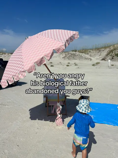 A beach scene under a bright blue sky. A baby in a blue shirt and patterned swim shorts is walking towards the shallow water. An adult is seated in a beach chair under a large pink and white checkered sun umbrella with a ruffled edge. A blue slip-n-slide is positioned on the sand. Text overlaid on the image poses a question about the father's abandonment.