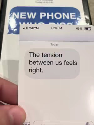 The image shows a phone screen displaying a text message conversation. The text bubble contains the message: "The tension between us feels right." Above the message, the phone shows the time as 4:20 PM and a battery life of 69%. In the background, there is a sign with the text "NEW PHONE WHO DIS?"