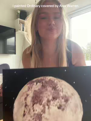 A woman is facing the camera, blowing a kiss. She has long blonde hair and is wearing a black top. Behind her, a painting of a full moon hangs on a wall. The background includes a window, shelves, books, and a television.