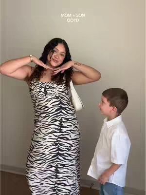 A woman and a young boy stand side-by-side in a well-lit indoor setting. The woman, on the left, models in a zebra print dress, accompanied by a white shoulder bag and gold jewelry. The boy, on the right, is dressed in a button-down white shirt and light-wash jeans. Text overlay at the top reads 'MOM + SON OOTD'.