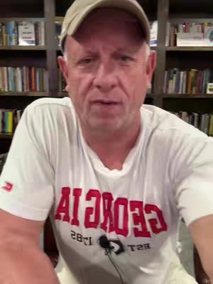 The cover image features a man sitting in front of a bookshelf. He is wearing a white t-shirt with red text, a beige cap, and a lavalier microphone. The background consists of a wooden bookshelf filled with books.