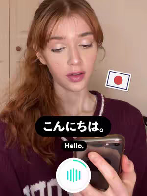 A young woman with long auburn hair is holding a phone in her hand, looking at the screen, and speaking. She is wearing a maroon sweater. There are Japanese characters and the English word 'Hello' overlaid on the image, in addition to a small Japanese flag sticker near her.