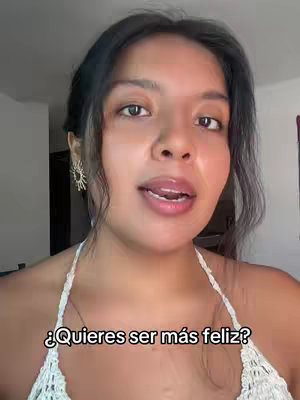 A woman with dark hair and a white top is centered in the frame, looking directly at the camera and speaking. The background is blurred, featuring what appears to be an interior space. Text overlay at the bottom reads: 'Quieres ser más feliz?'