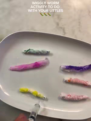 A top-down shot of a white platter holding several colorful, wiggly worm-like creations. The worms are made of colorful materials and placed on the plate, alongside a syringe. The background is a light, neutral tone evoking a sense of cleanliness and simplicity.