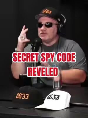 A man wearing sunglasses, a black cap with 'gg33' on it, and a headset is positioned in a dark area. The background is indistinct. In front of him are two hats: one black with gold text and another white with black text. The video overlayed text reads 'SECRET SPY CODE REVEALED'. A microphone is positioned near the man's face.
