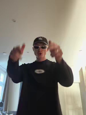 A man is standing in front of a camera with his arms raised and index fingers pointed towards the camera. He wears a black cap with a white logo, white sunglasses, and a black sweatshirt. The background is a plain ceiling and some light. The overall composition is simple, with the focus on the man's expression and gesture.