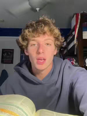 A young man with curly blonde hair looks directly at the camera, his mouth slightly open as if speaking. He's wearing a dark blue hoodie, and the background is a blurred interior with a ceiling fan and glimpses of shelves and books. An open book is visible in the foreground.