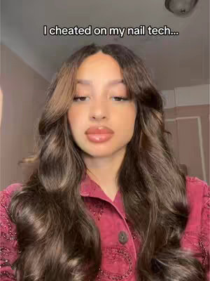 A young woman with long, wavy brown hair is centered in the frame. She has full, glossy lips and is wearing a reddish-purple top. Text overlaid above the woman reads 'I cheated on my nail tech...' The background is softly lit with neutral tones, and a light fixture is visible at the top.