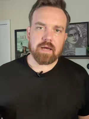 A man with a beard and dark hair is centered in the frame. He wears a black t-shirt and speaks directly to the camera. The background includes a wall and framed pictures. The lighting is natural, and the composition is a medium shot.