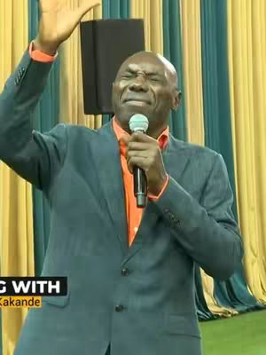 A man in a gray suit and orange shirt is holding a microphone and speaking. The background features a yellow and blue pattern, and some text is added.