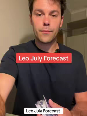 A man with dark hair, wearing a black t-shirt, is the central figure. He is looking at the camera with a neutral expression. Above him, a red rectangle reads 'Leo July Forecast', overlaid on the image in white text, and below him, a rectangle with the same text, but in smaller font. His hands hold a deck of cards in front of him. The lighting is fairly even, and the background is out of focus. The composition is a medium shot framing the subject from the chest up.