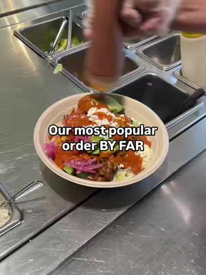 A top-down shot shows a beige bowl filled with various food items. The bowl is centered against a stainless steel counter, with storage bins visible in the background. A person is pouring a reddish sauce over the bowl from a clear bottle, and text overlays the bowl.