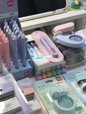 A well-lit, top-down shot of various stationery items arranged on a white surface. The items include pens, a pink and blue stapler, tape, and a utility knife, all neatly displayed. The color palette is pastel with a focus on blues, pinks, and whites. The composition is clean and organized, with items partially visible, contributing to an unboxing or product display feel.