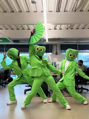 Three figures in green suits, each wearing a Duolingo owl mask, strike dynamic poses in a well-lit, modern office space. The central figure holds up a green fan. The scene conveys a playful, promotional vibe associated with the language-learning app Duolingo.