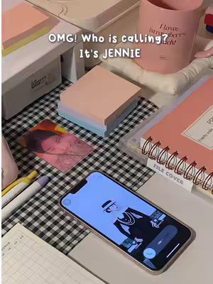 The image is a well-lit, close-up shot of a desk setup with a variety of items. The foreground features an Apple iPhone with a call screen showing a black and white image of the celebrity Jennie. Adjacent to it, there are a stack of peach-colored post-it notes, a pink mug with "I love strawberry lunch!" printed on it, a pink spiral-bound notebook, pens, a photocards featuring Jennie, and a decorative white box. The background consists of a checkered black and white tablecloth. The overall composition has a warm, inviting feel.